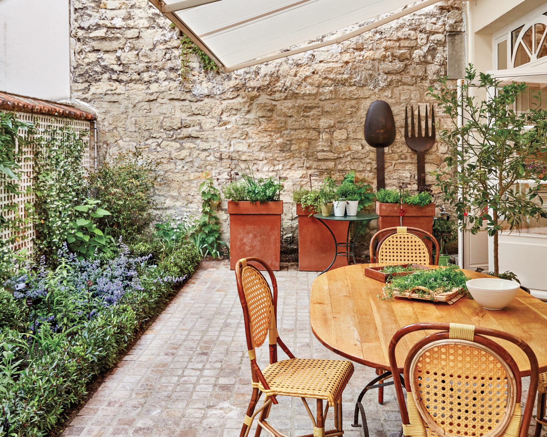 Charming outdoor patio with wooden table, wicker chairs, potted plants, and herbs against a rustic stone wall background.
