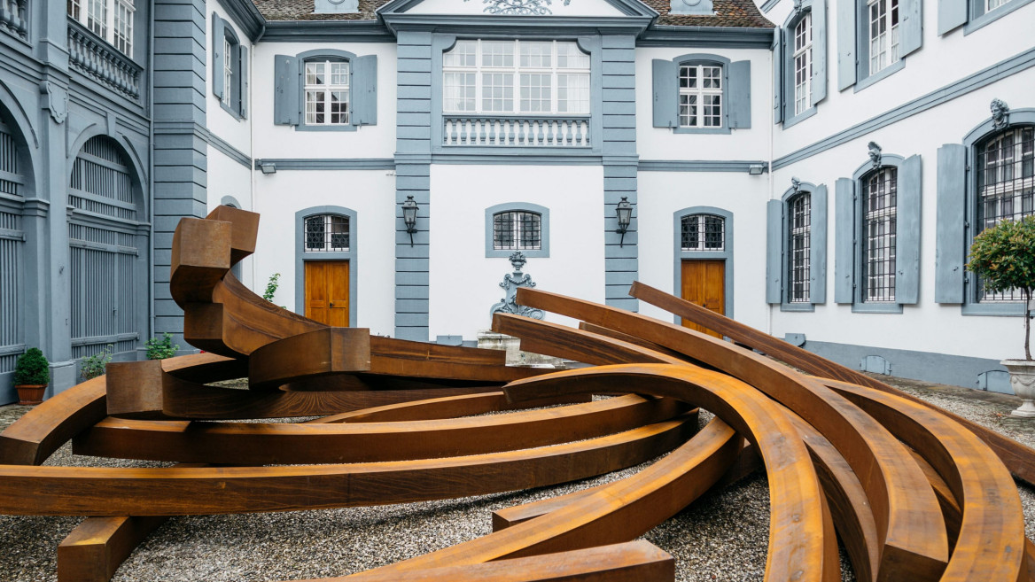 Contemporary metal sculpture with curved beams in courtyard of historic blue and white building with large windows.