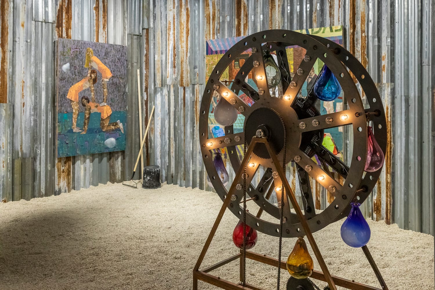 Ferris wheel-like sculpture with colorful bulbs in a rustic gallery, corrugated metal walls, and abstract paintings in the background.