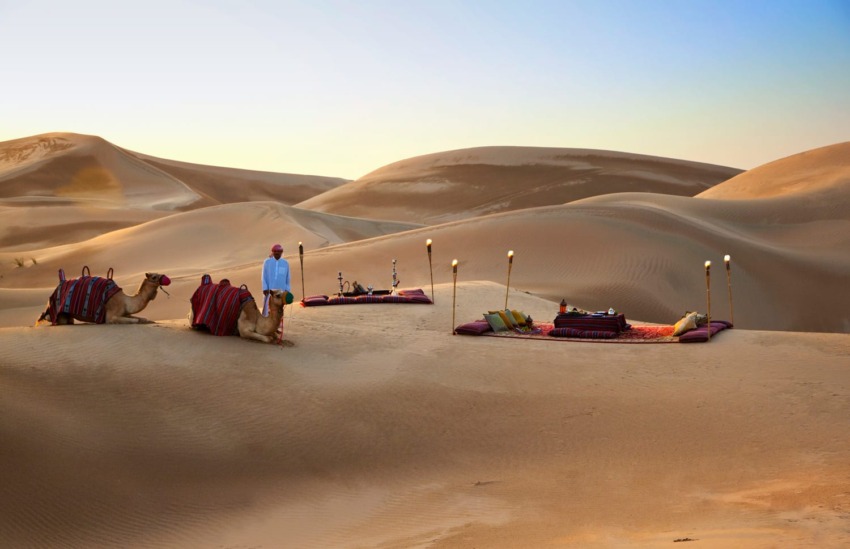 Man standing next to camels and a cozy seating area set up on sand dunes at sunset.