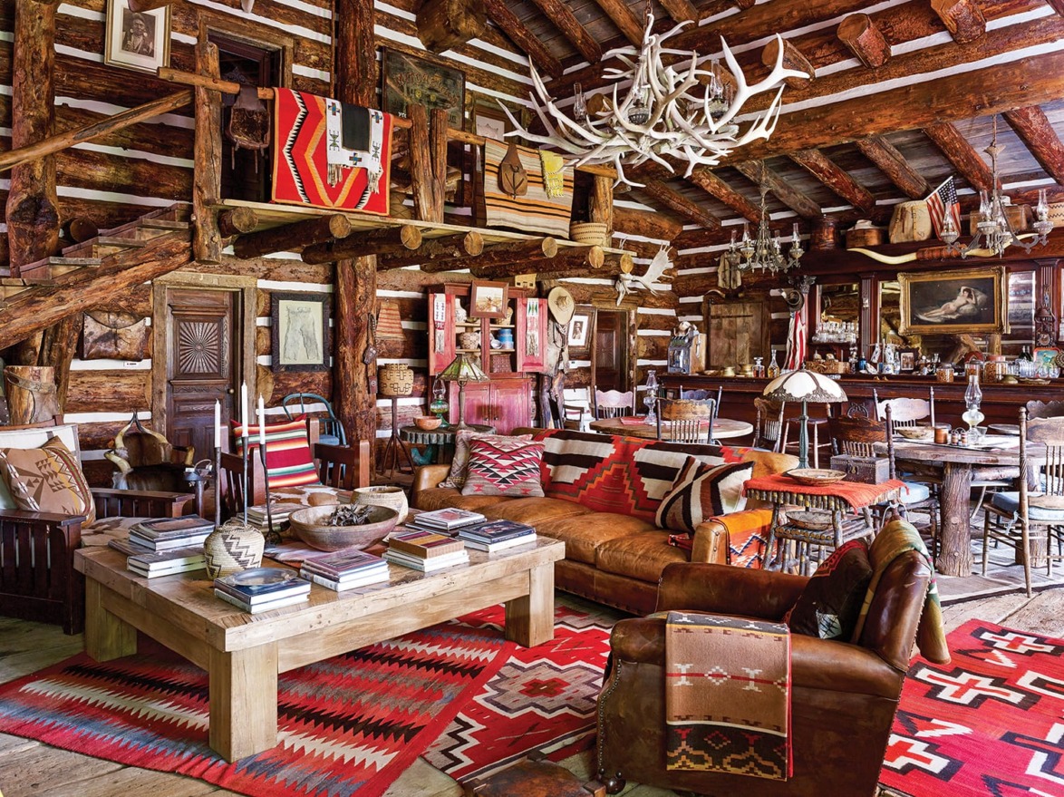 Rustic log cabin interior with wooden beams, antler chandelier, leather furniture, colorful textiles, and books on a table.