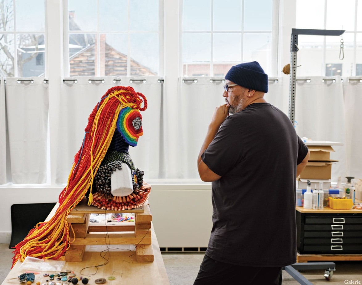 Artist contemplating a colorful sculpture with long yarn hair in a studio setting with large windows and various art supplies.