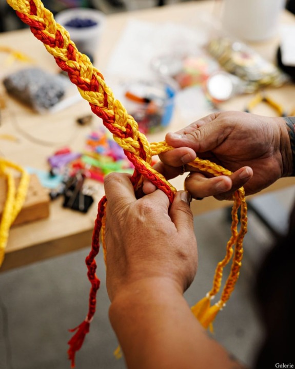 Hands braiding colorful red and yellow fibers on a table with crafting materials in the background.