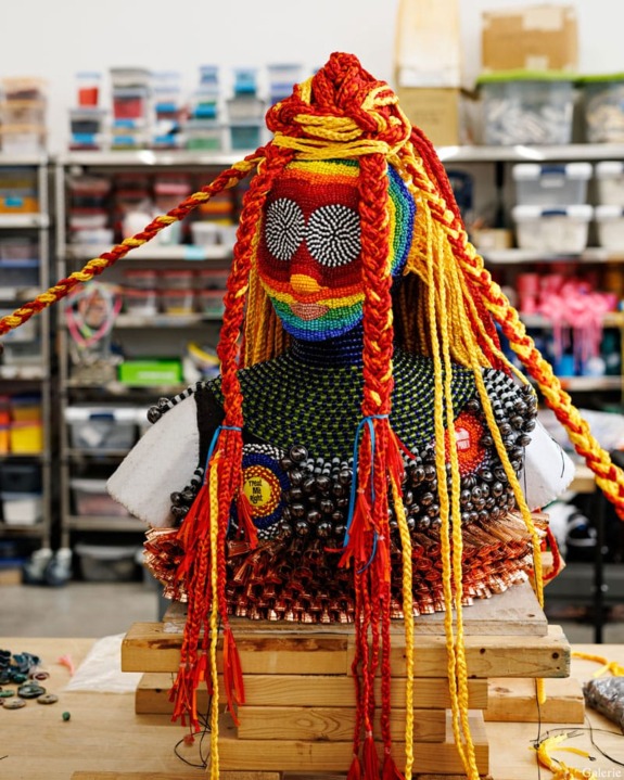 Colorful beaded sculpture with braided yarn hair on a table in an art studio filled with supplies.