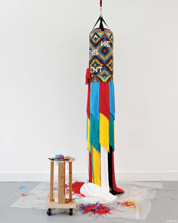 Colorful fringed punching bag art piece with geometric patterns, paint splatters on floor, and small table with art supplies nearby