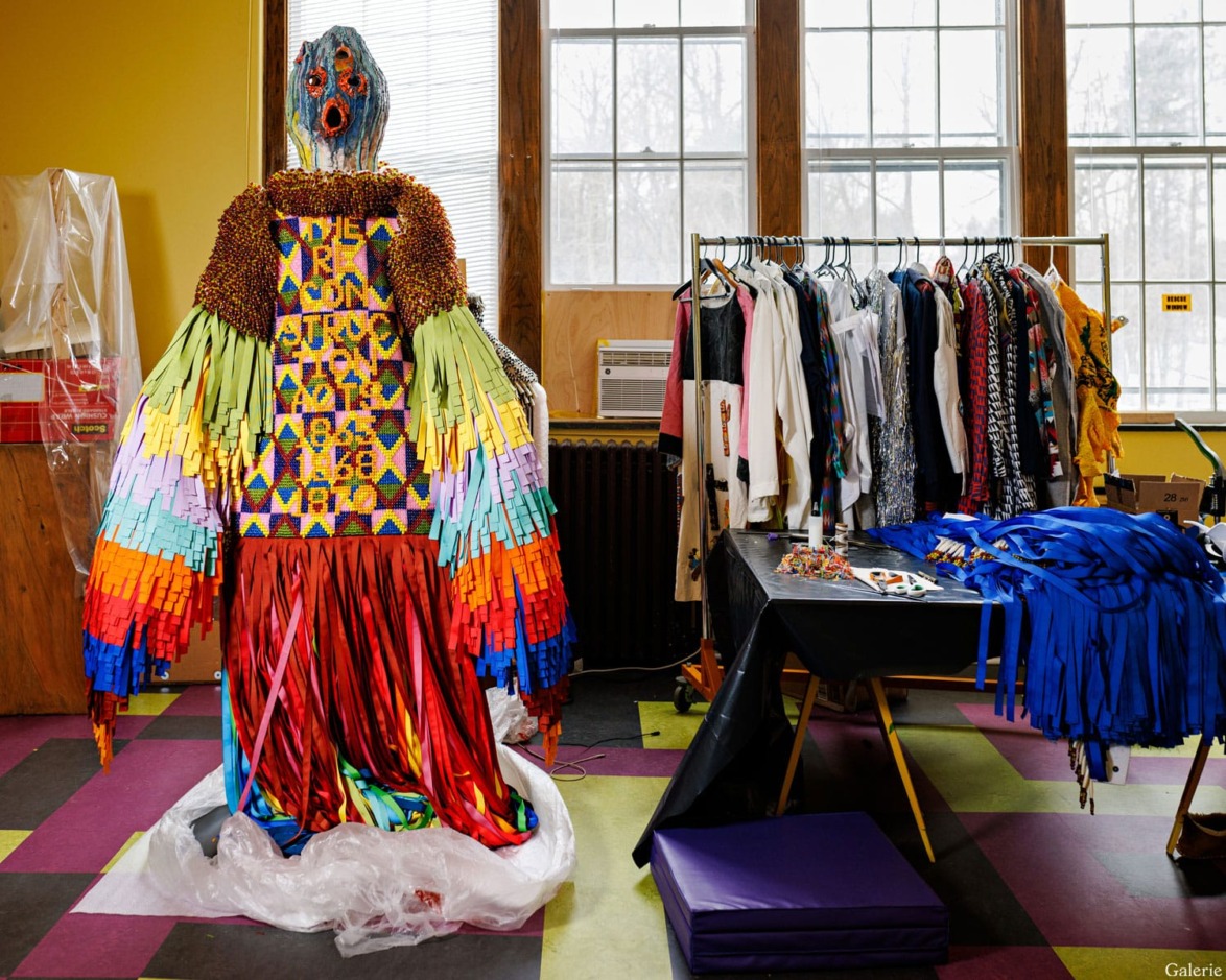 Colorful costume with fringe details displayed in an art studio, surrounded by clothing racks and colorful fabric.