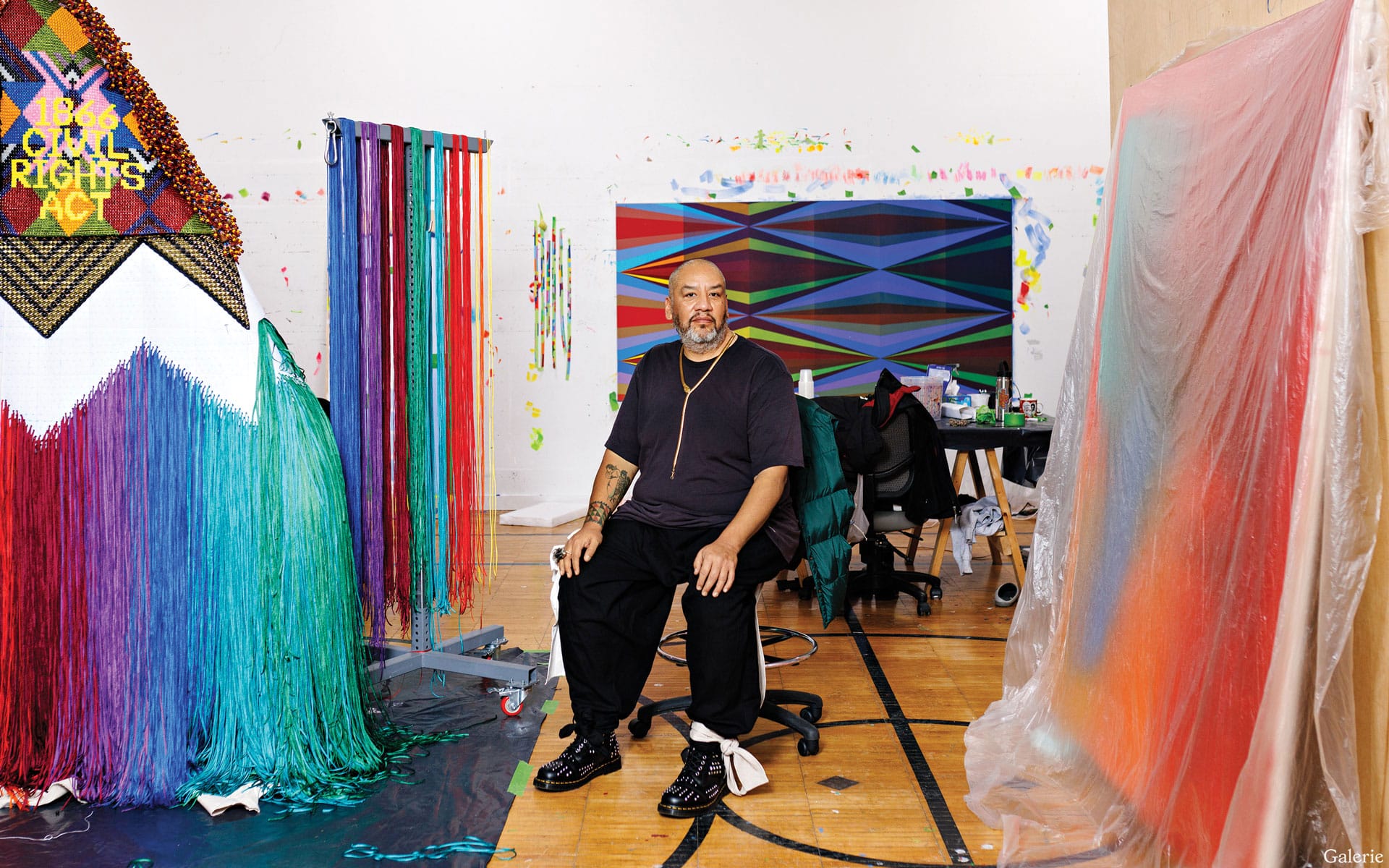 Artist sitting in a colorful studio with vibrant, abstract artwork and materials surrounding him