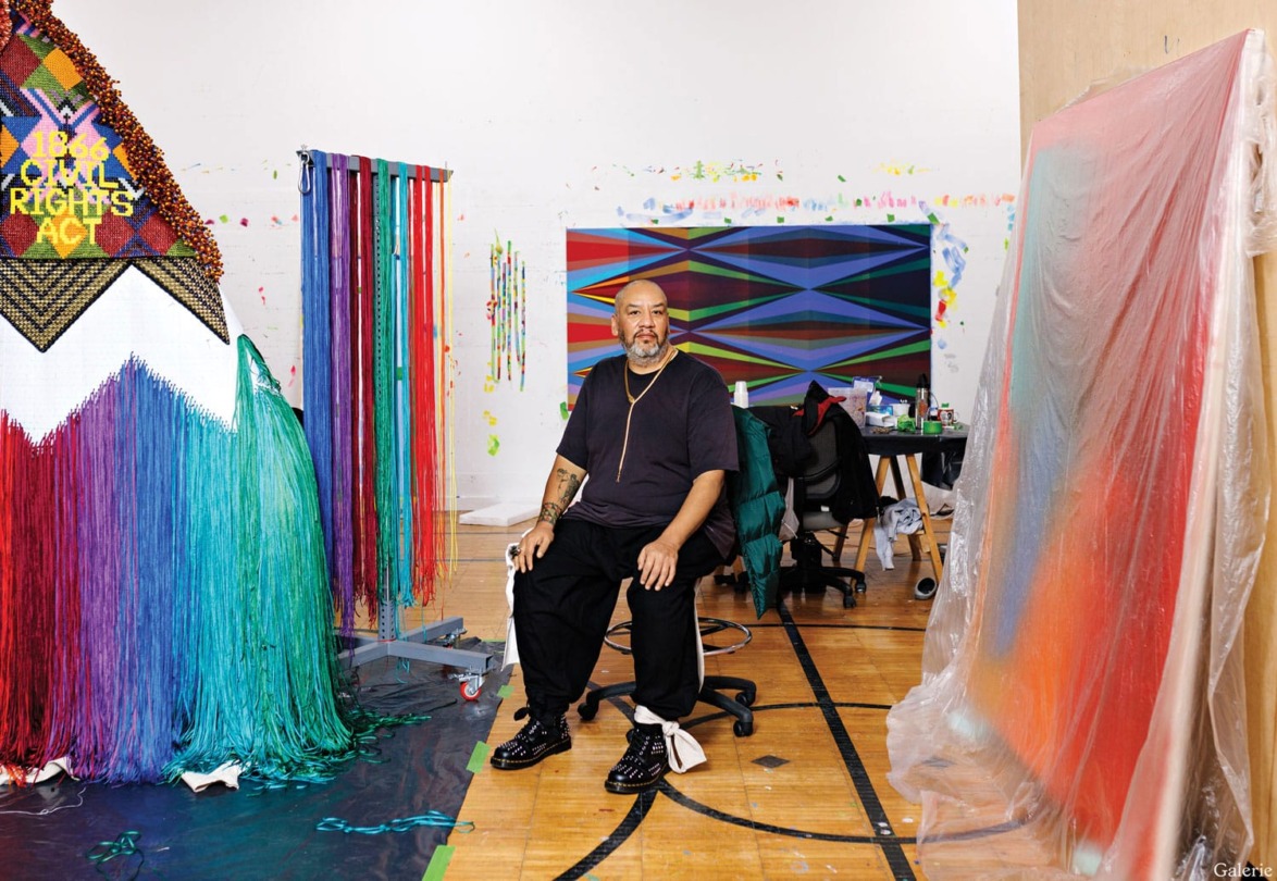 Artist sitting in a colorful studio surrounded by vibrant textile art and abstract paintings.