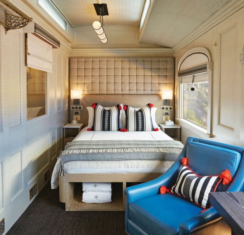Luxury train cabin with elegant decor, a plush bed, blue armchair, and natural light streaming through large windows.
