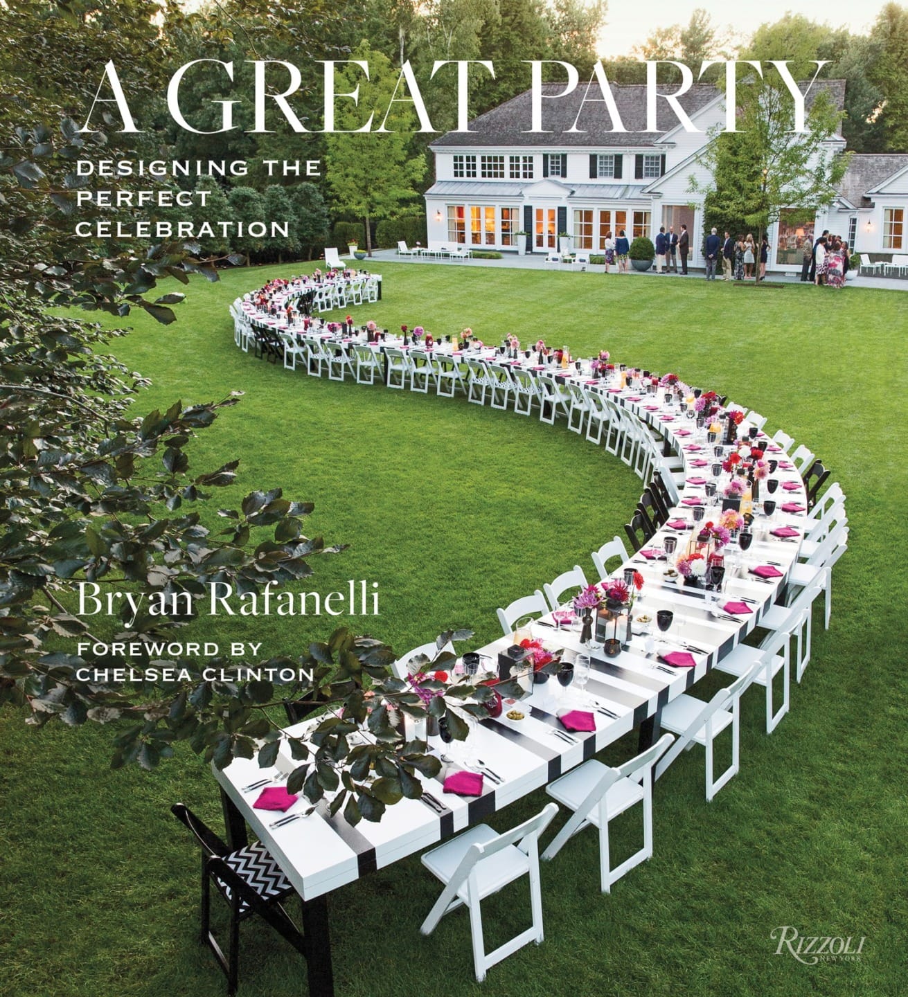 A long, curved outdoor dining table set with pink and white decorations on a lawn in front of a large house.