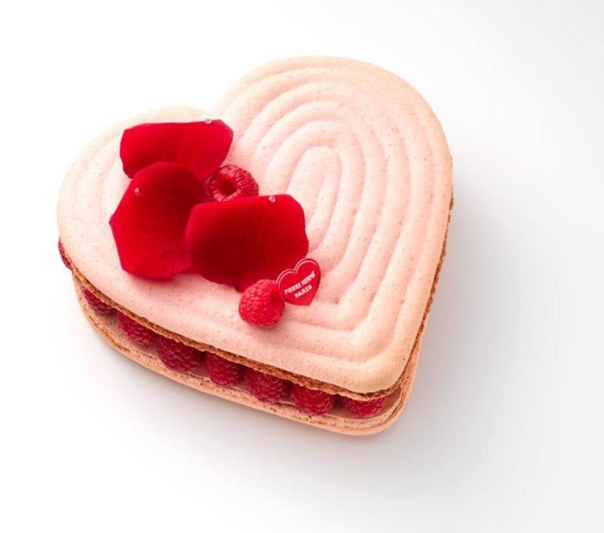 Heart-shaped pink cake with raspberries and red rose petals on top, placed on a white background.