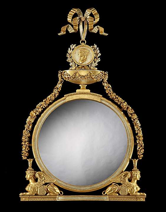 Ornate round mirror with gold frame, ribbon at top, floral garlands, and decorative sphinx figures on sides.
