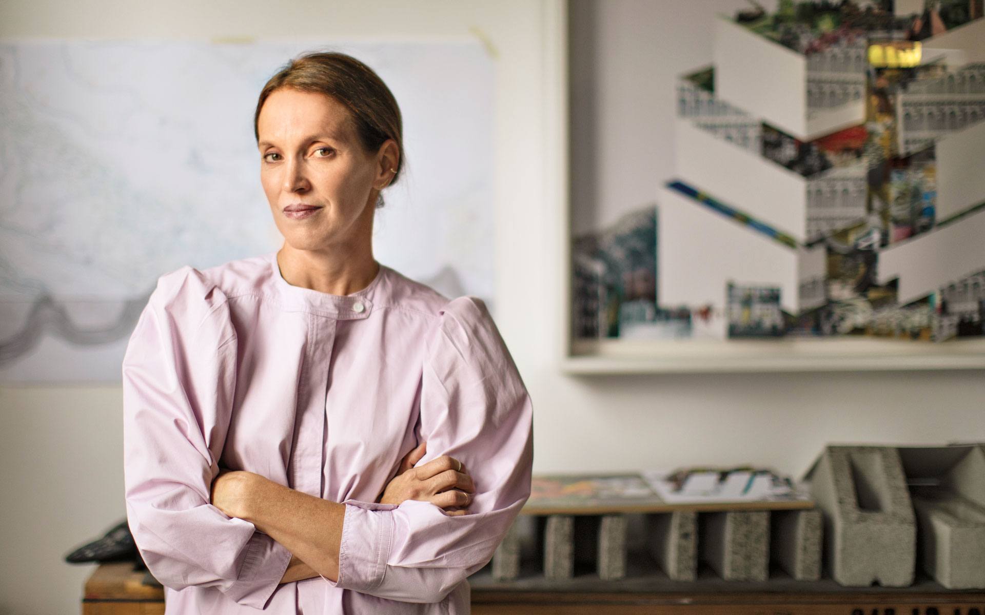 Woman in a light pink blouse stands confidently with arms crossed in a room with architectural designs in the background.