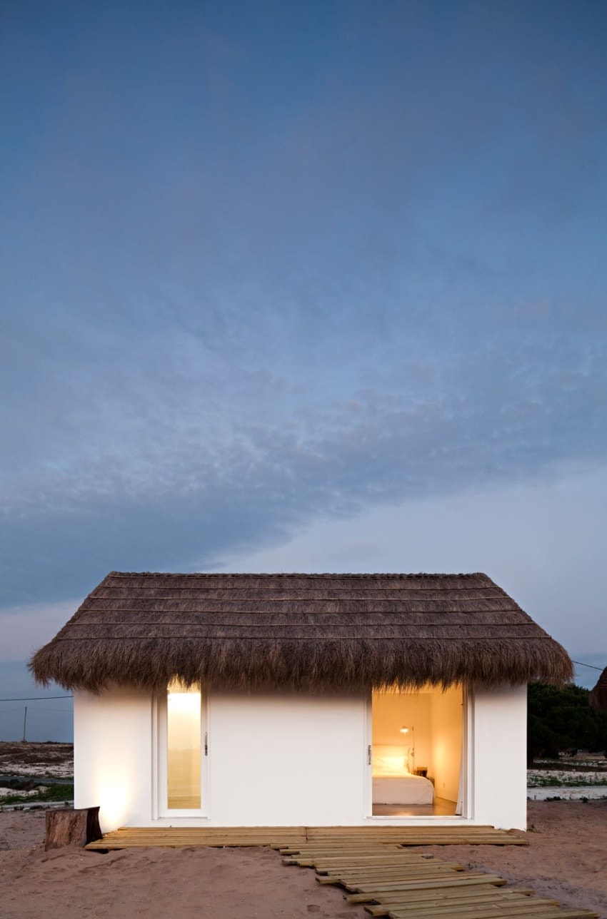 Small white beach house with thatched roof, warm interior lighting at dusk, wooden path leading to entrance.