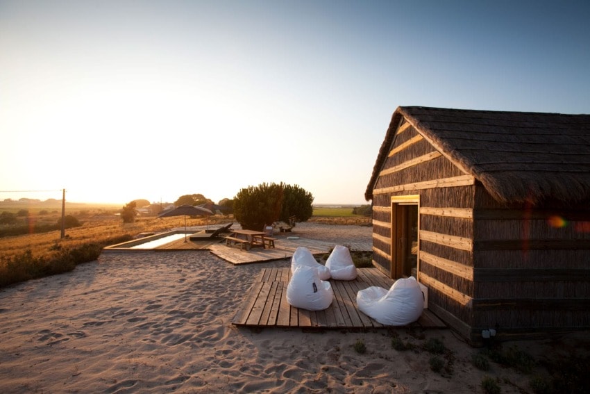 Rustic cabin with straw roof at sunrise, surrounded by sandy landscape, wooden deck, and white bean bags.