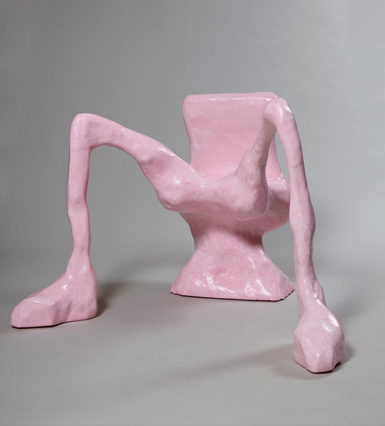 Abstract pink sculpture resembling a chair with elongated, irregular legs on a gray background.