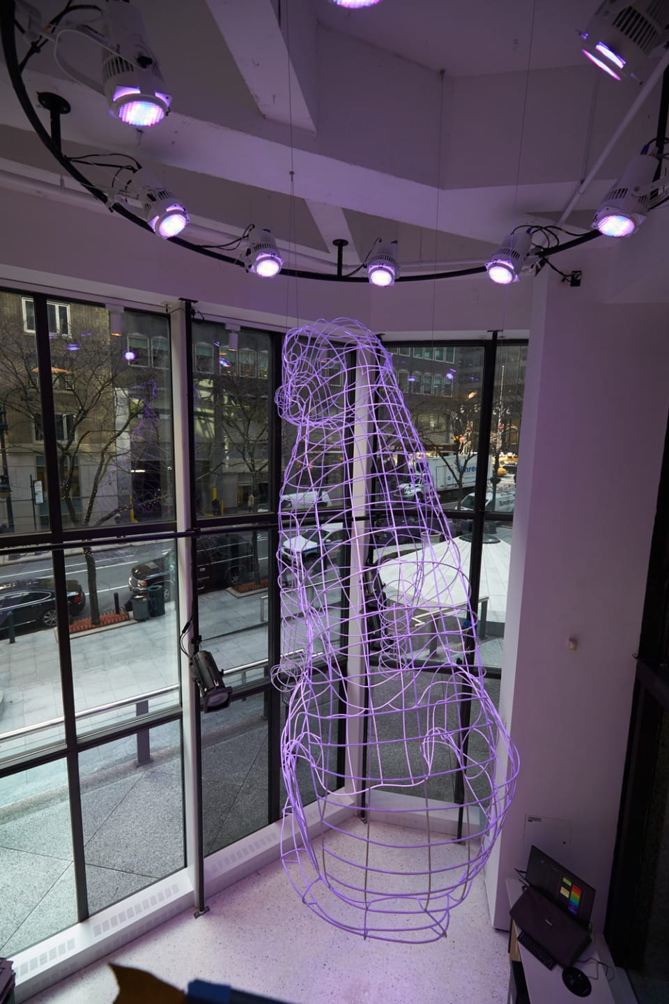 Purple wireframe sculpture suspended from ceiling in modern building with floor-to-ceiling windows and street view outside.