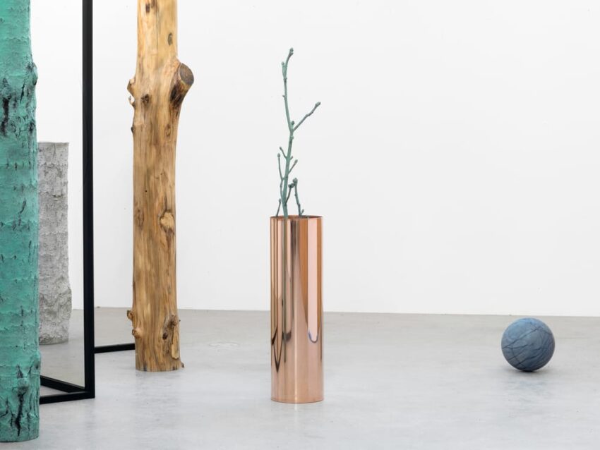 Modern art display with a copper vase holding twigs, blue sphere, and wooden sculptures in a minimalist gallery setting.