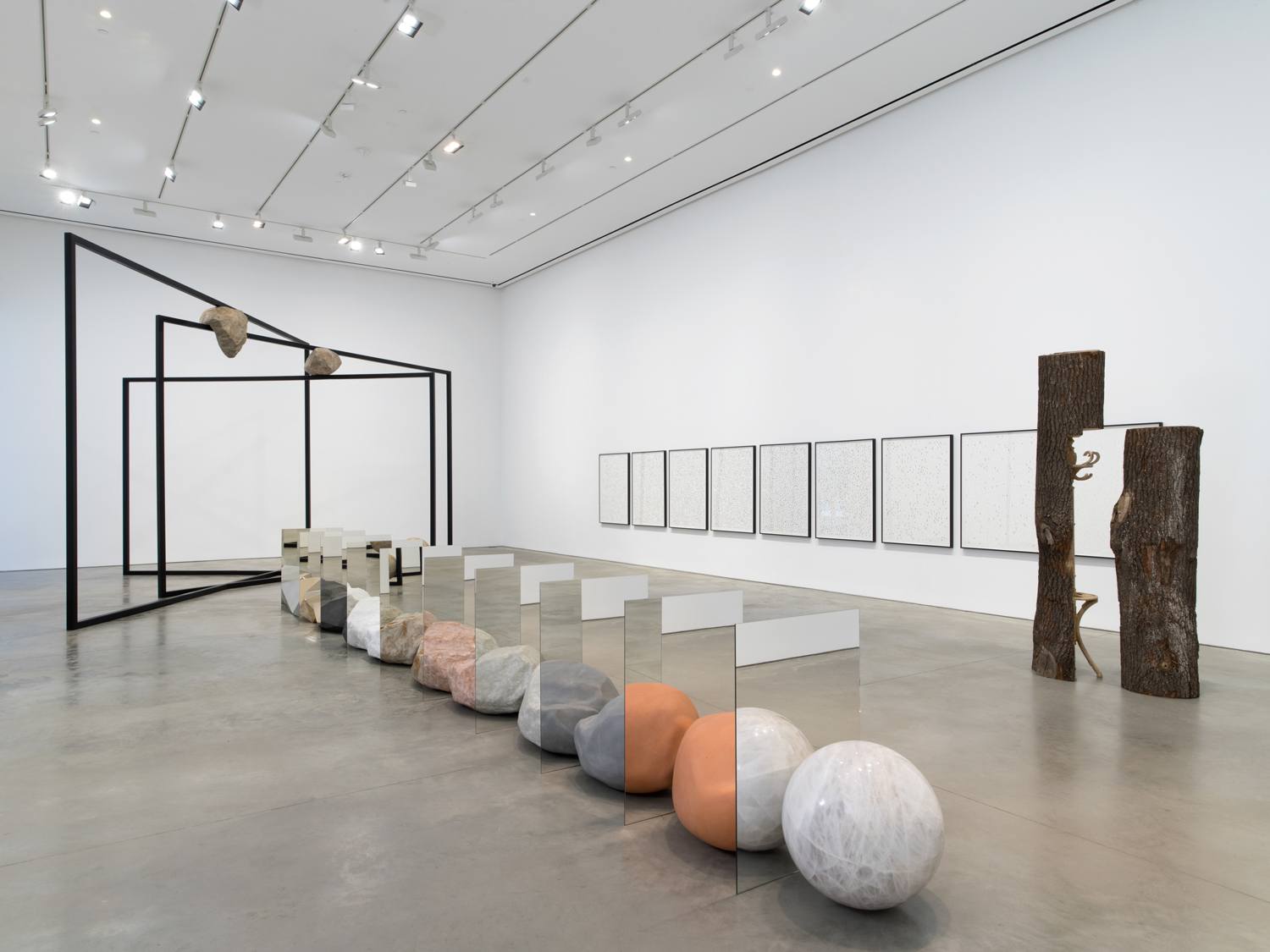 Contemporary art installation with geometric sculptures, mirrors, spheres, and framed artworks in a gallery setting.