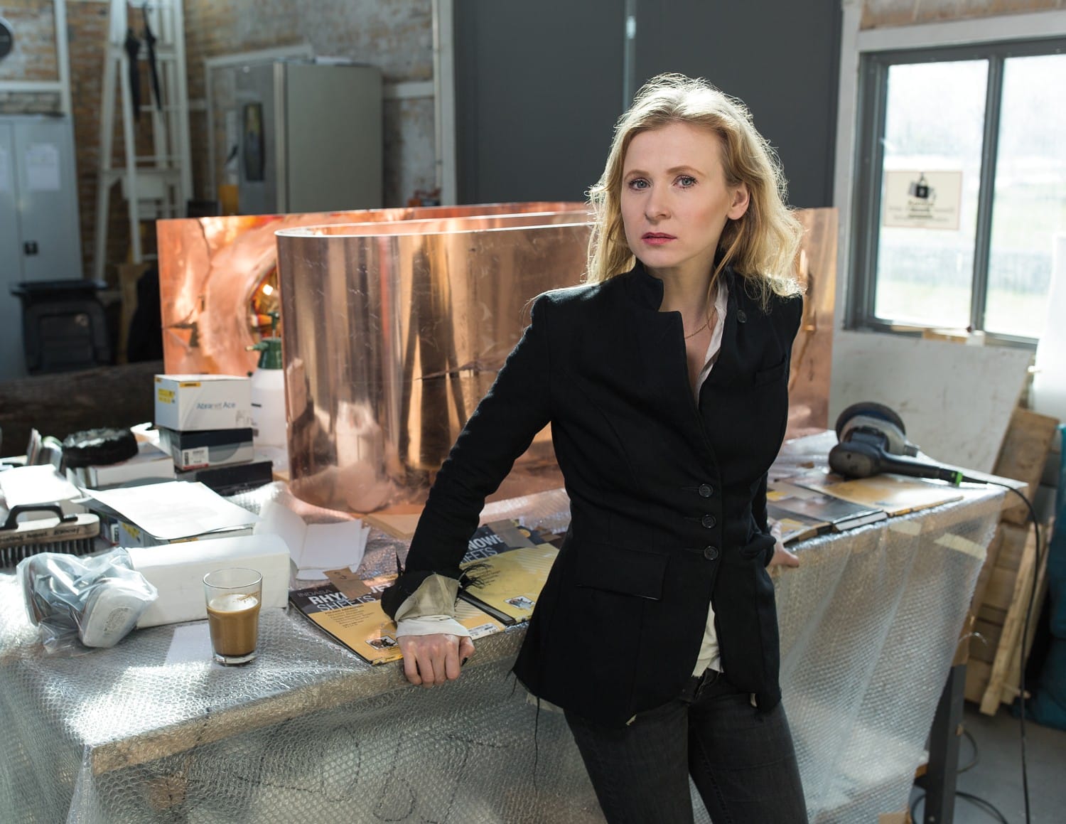 Person in a black jacket stands in an industrial setting with copper in the background.