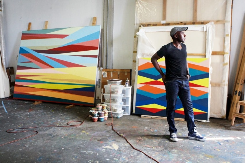 Artist standing in a studio with colorful abstract paintings leaning against the wall and art supplies scattered on the floor.