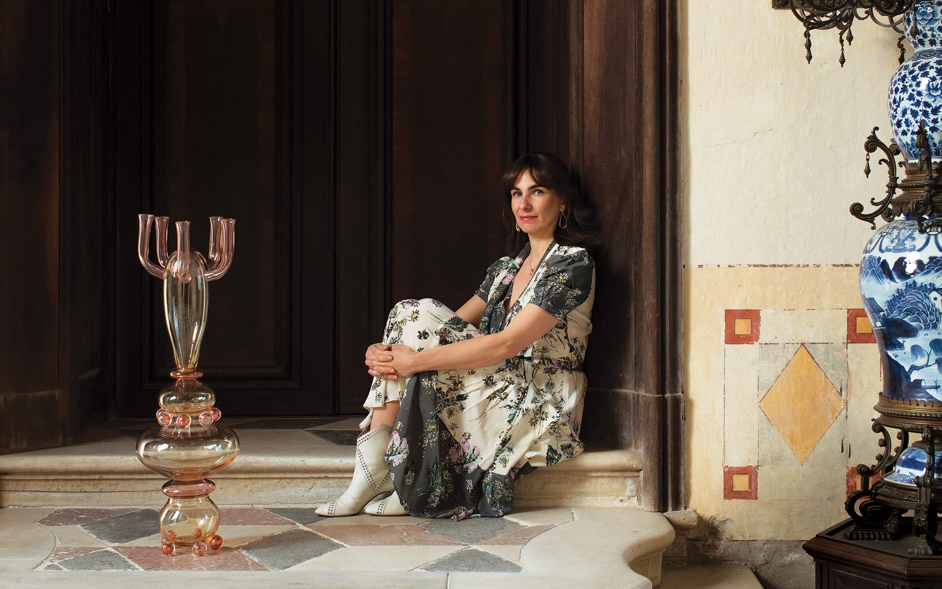 Woman in a floral dress sits on stone steps indoors next to glass sculpture and ornate vases.