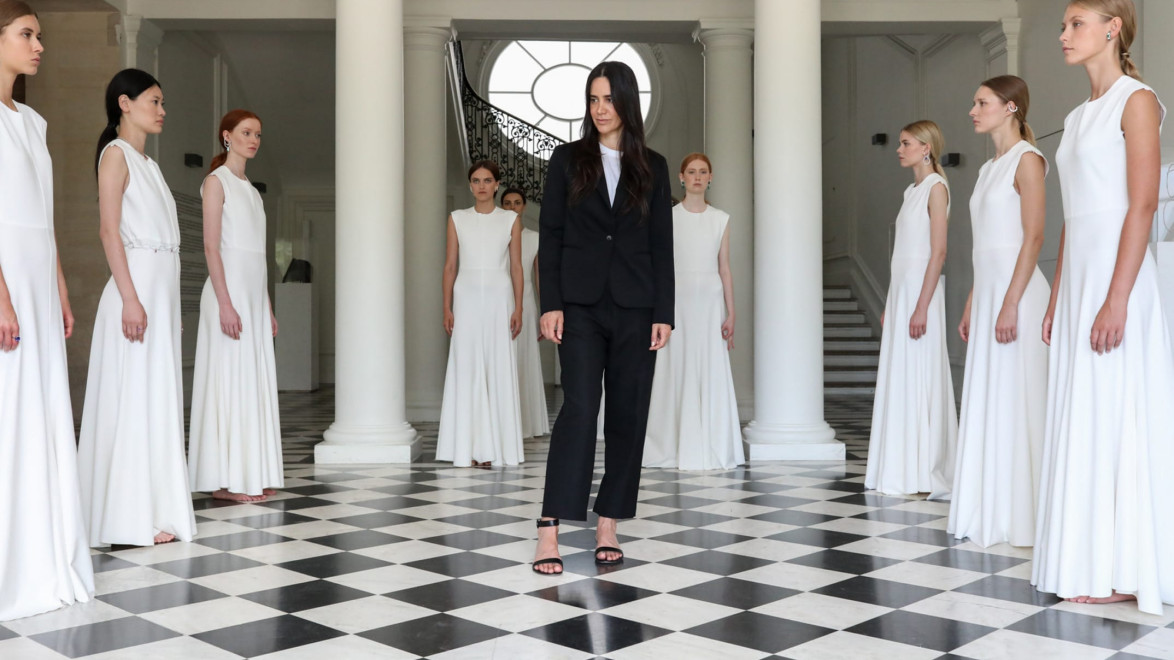A woman in a black suit stands in a marble-floored room, surrounded by women in white dresses, with columns in the background.