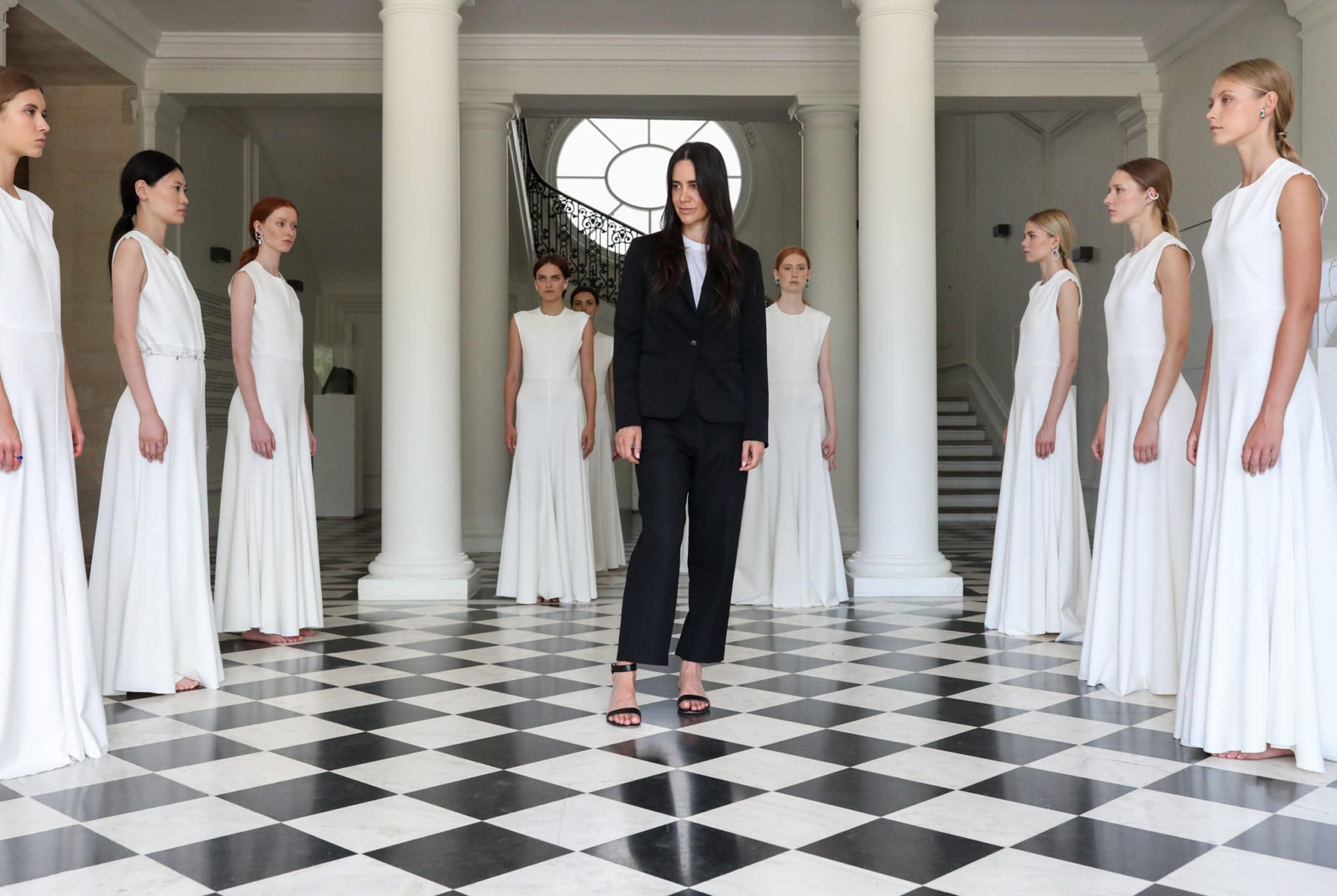A woman in a black suit stands in a marble-floored room, surrounded by women in white dresses, with columns in the background.