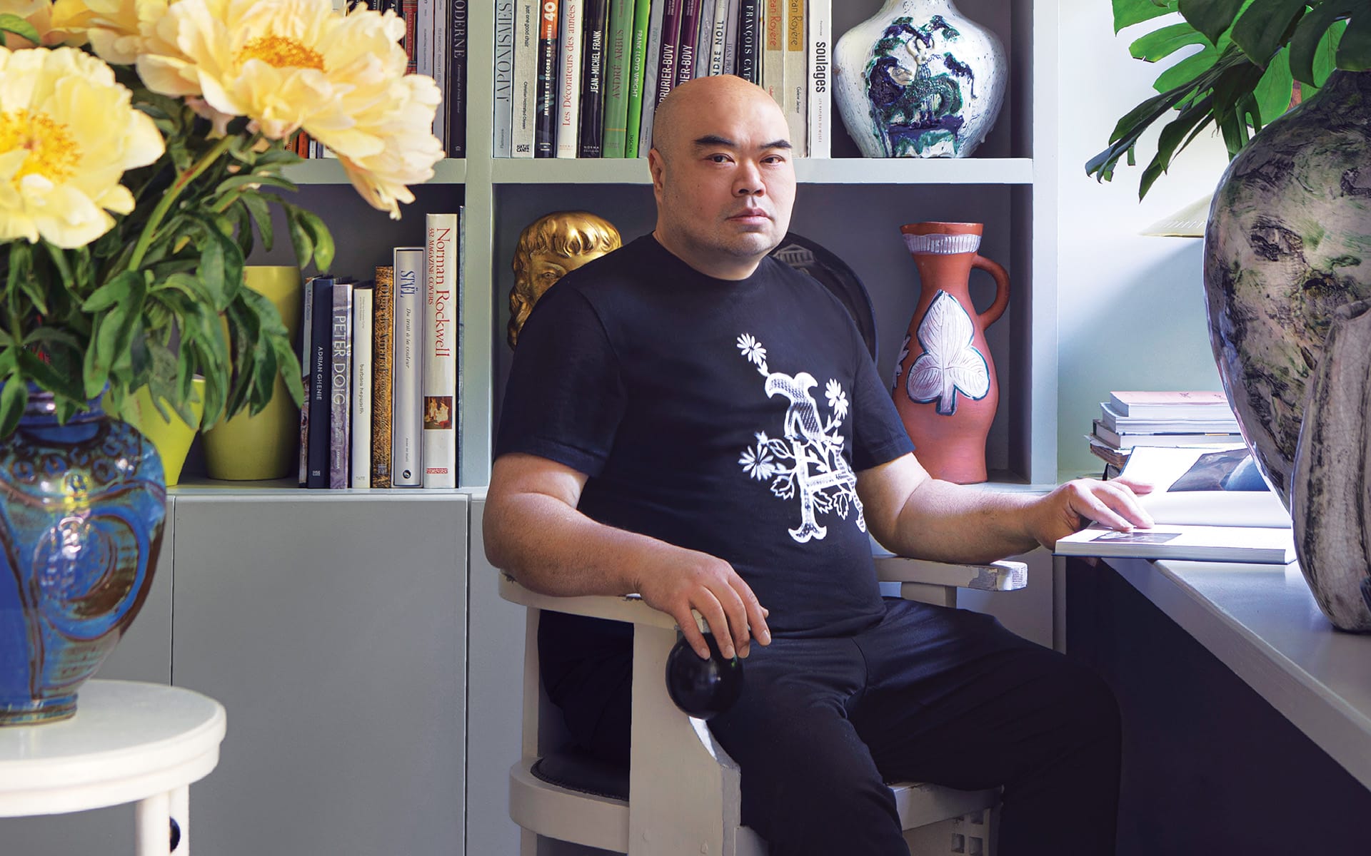 Person seated in a library with shelves of books and decorative vases, looking at the camera with a neutral expression.