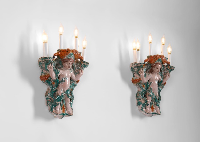 Two ceramic wall sconces with candle-shaped bulbs and intricate detailing of human figures and foliage on a plain background.
