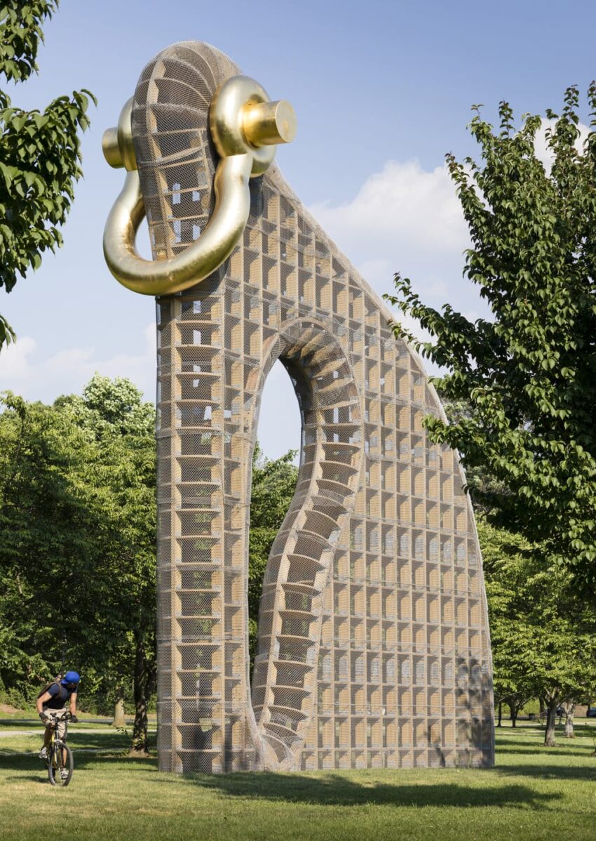 Giant sculpture resembling an earring with golden accents in a park setting, surrounded by trees and a cyclist nearby.