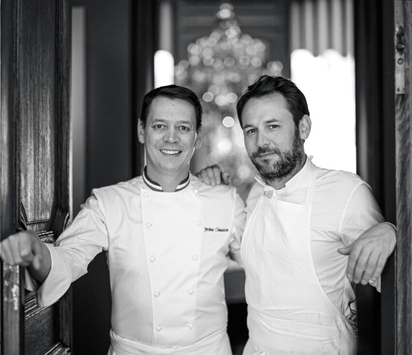 Two chefs in white jackets standing in a doorway, one with arms crossed, smiling, and the other with hands on the door frame.