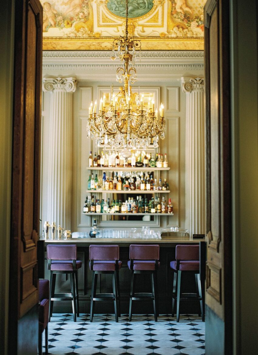 Elegant bar with a luxurious chandelier, high ceiling, and rows of colorful bottles on display behind purple bar stools.