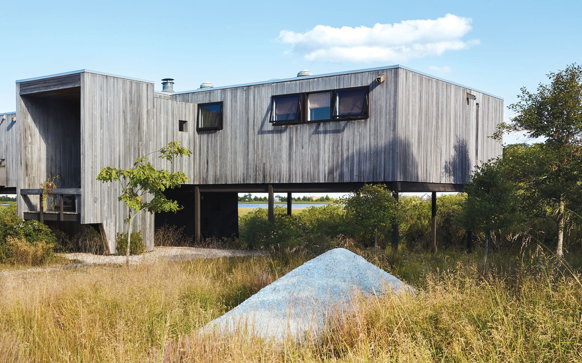 Modern elevated wooden house among tall grasses and trees, with a small blue gravel mound in the foreground under a blue sky.