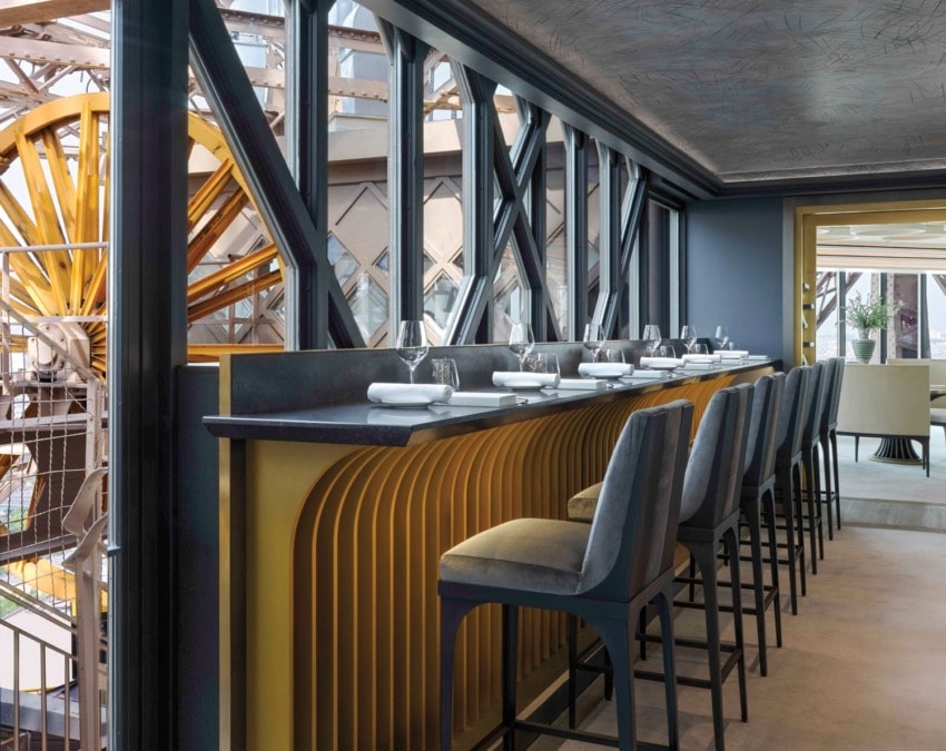 Modern restaurant interior with tall bar chairs and set tables near large windows, featuring a stylish decor and spacious layout.