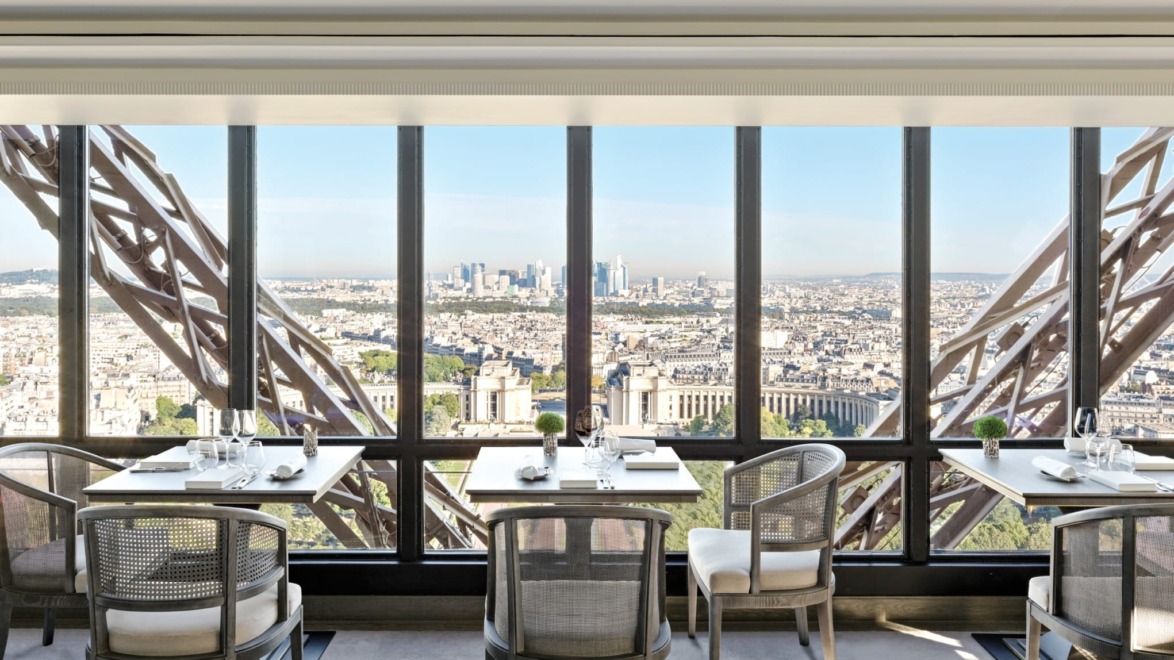 Dining area with tables and chairs offering a panoramic view of a cityscape through large windows in an elevated setting.