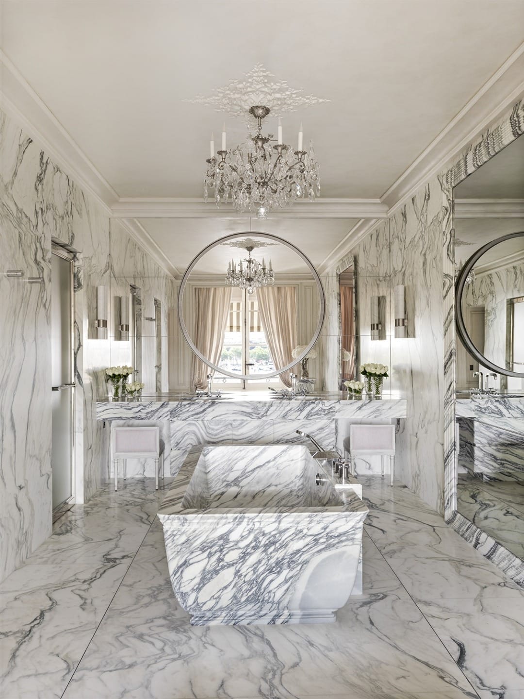 Luxurious marble bathroom with freestanding tub, large mirrors, elegant chandelier, and tall windows with curtains.