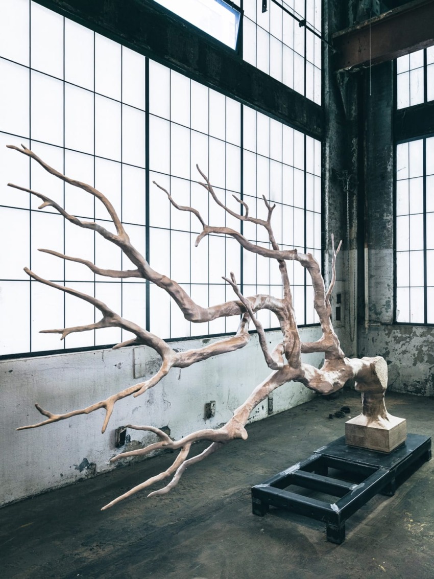 Sculptural artwork of a tree branch displayed in an industrial-style gallery with large windows and concrete floors.