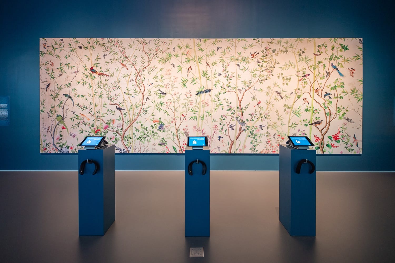 Interactive museum exhibit with three touchscreen displays in front of a large decorative panel featuring colorful birds and foliage.