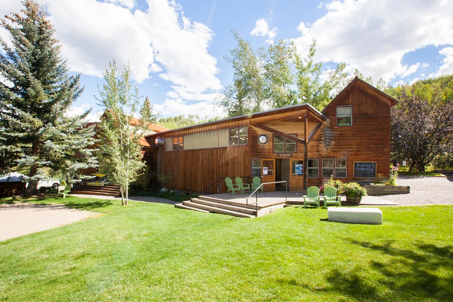 Wooden building with large windows surrounded by green lawn, trees, and Adirondack chairs under a partly cloudy sky.