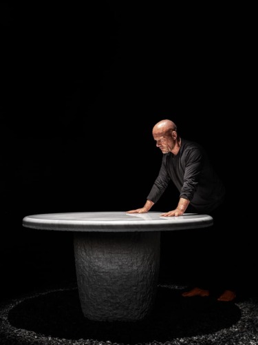 Man leaning on a round stone table in a dimly lit setting, wearing a black shirt, with dramatic lighting.