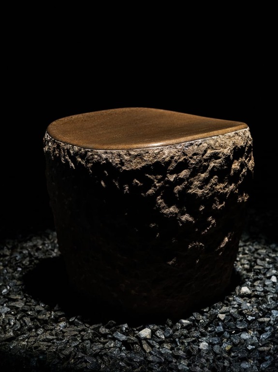 Dark stone sculpture with a smooth top surface, surrounded by small black pebbles.
