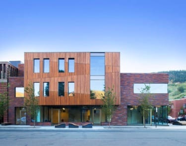 Modern building with wood and glass facade, large windows, and small trees lining the street under a clear blue sky.