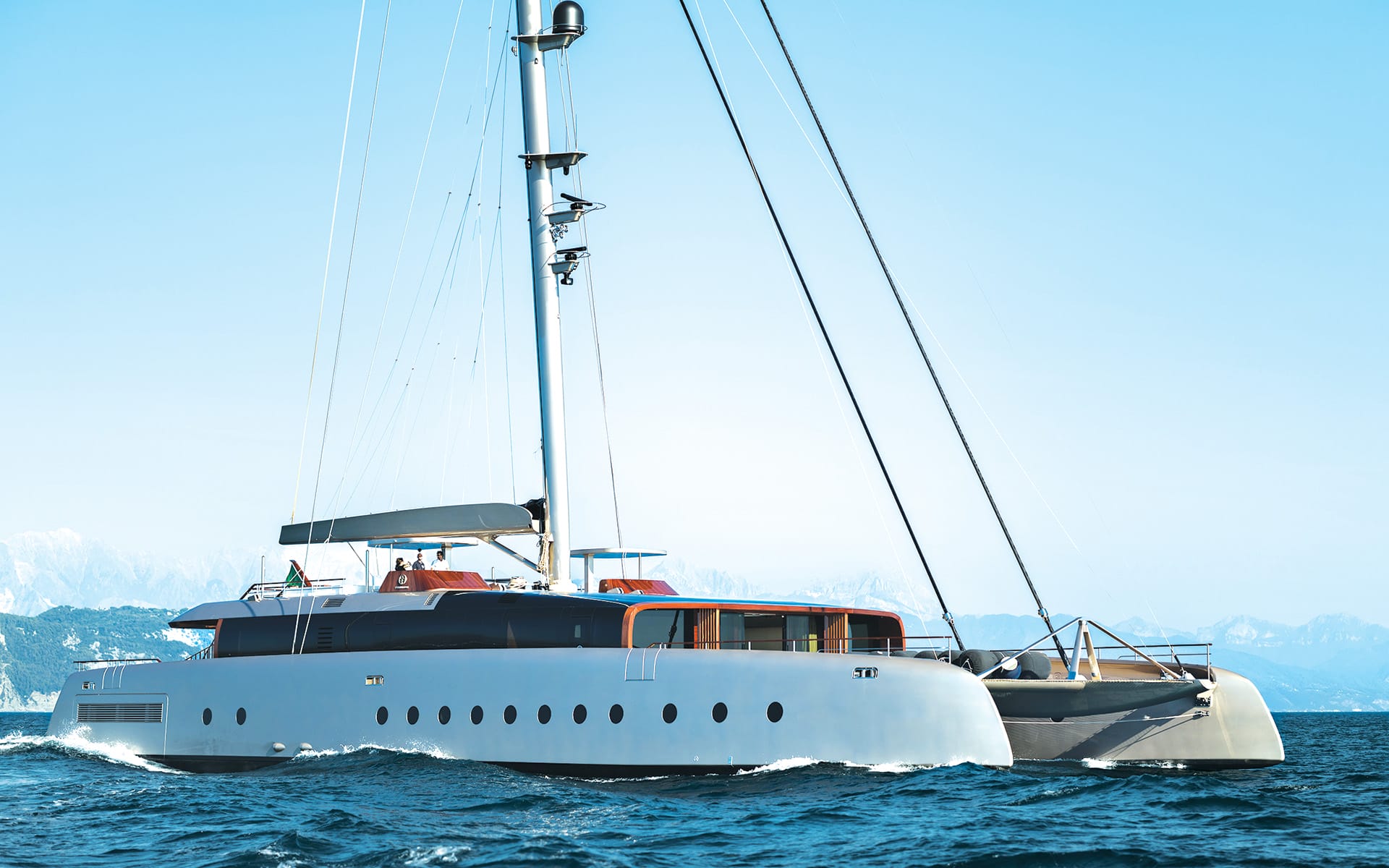 Large white sailing yacht navigating calm blue waters with mountains in the background.