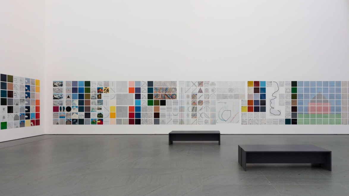 Art gallery with colorful tiled wall display and two black benches on a gray floor.