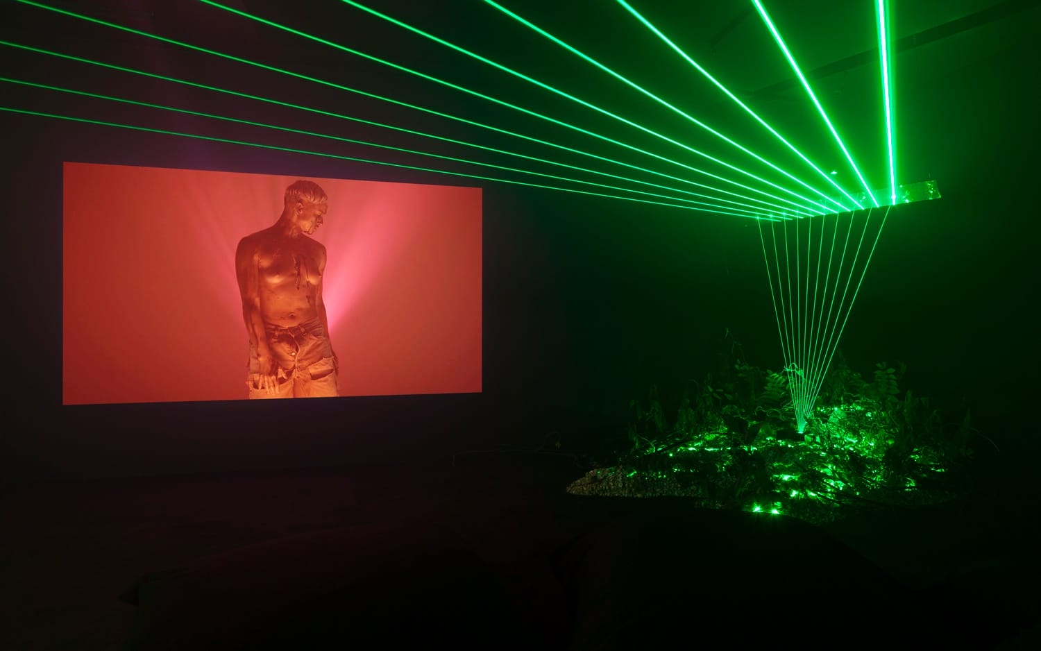 Art installation with green lasers and a screen displaying a video of a standing man in a dimly lit room.