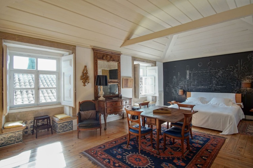 Spacious bedroom with wooden floor, vintage furniture, large bed, and chalkboard wall art, featuring two bright windows.
