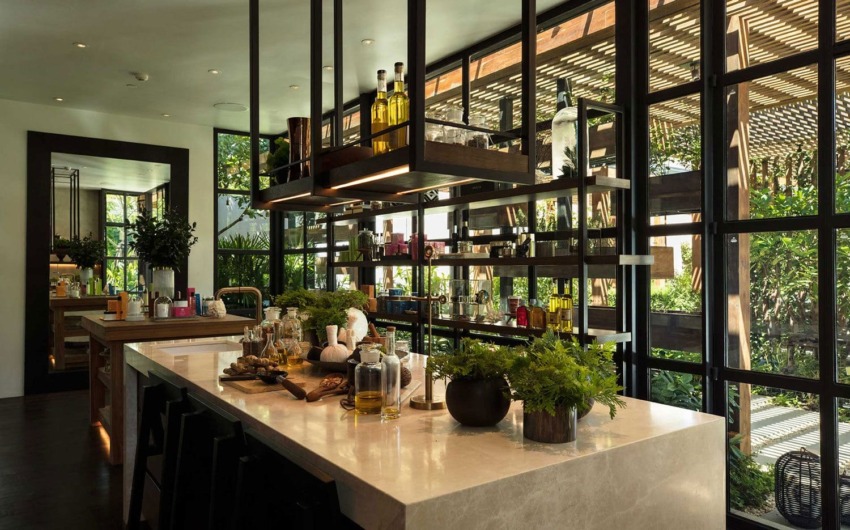 Modern kitchen with large island, bar stools, hanging shelves, and greenery near large windows letting in natural light.
