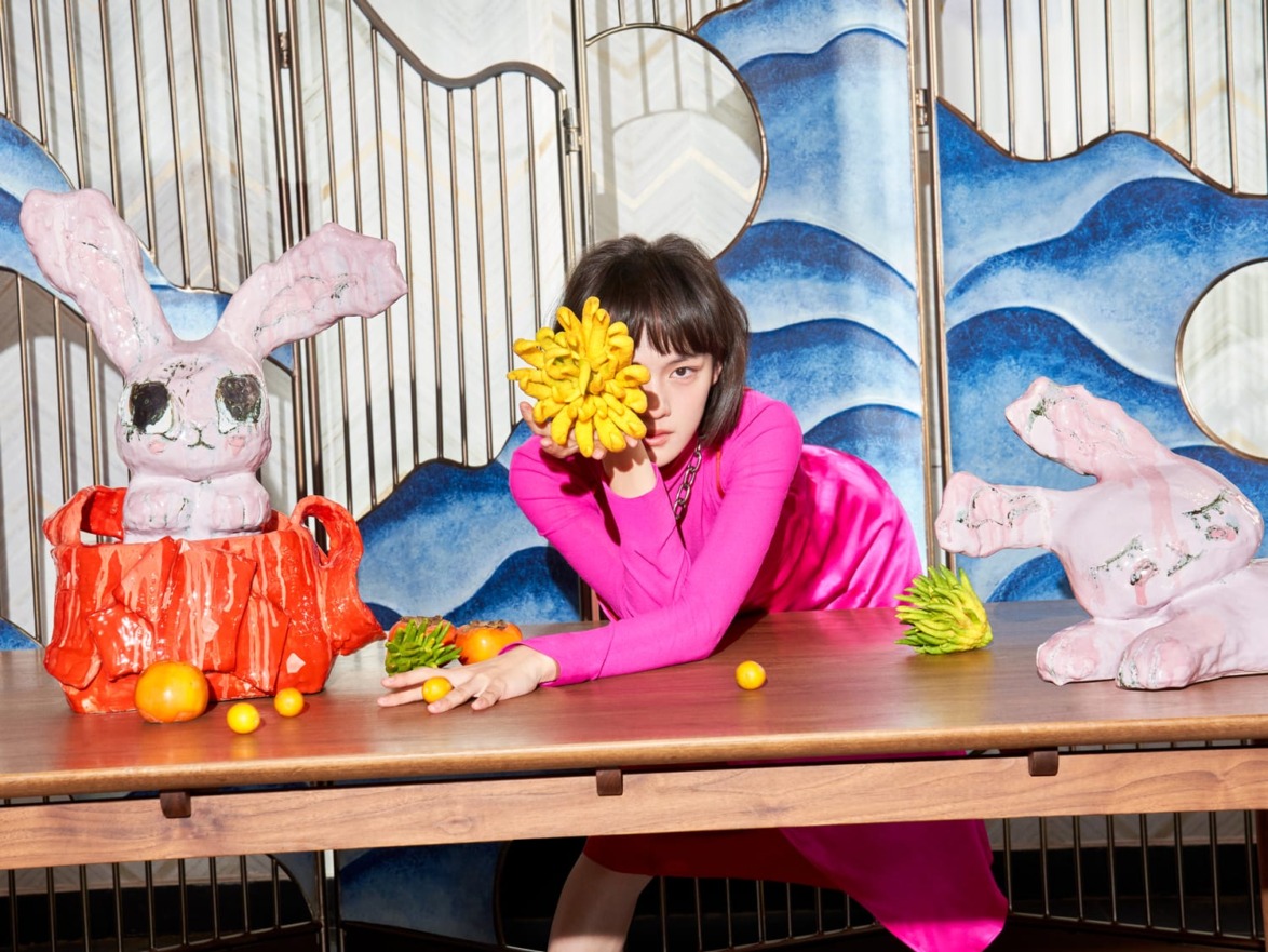 Woman in pink dress posing with artistic sculptures and tropical fruits on a table.