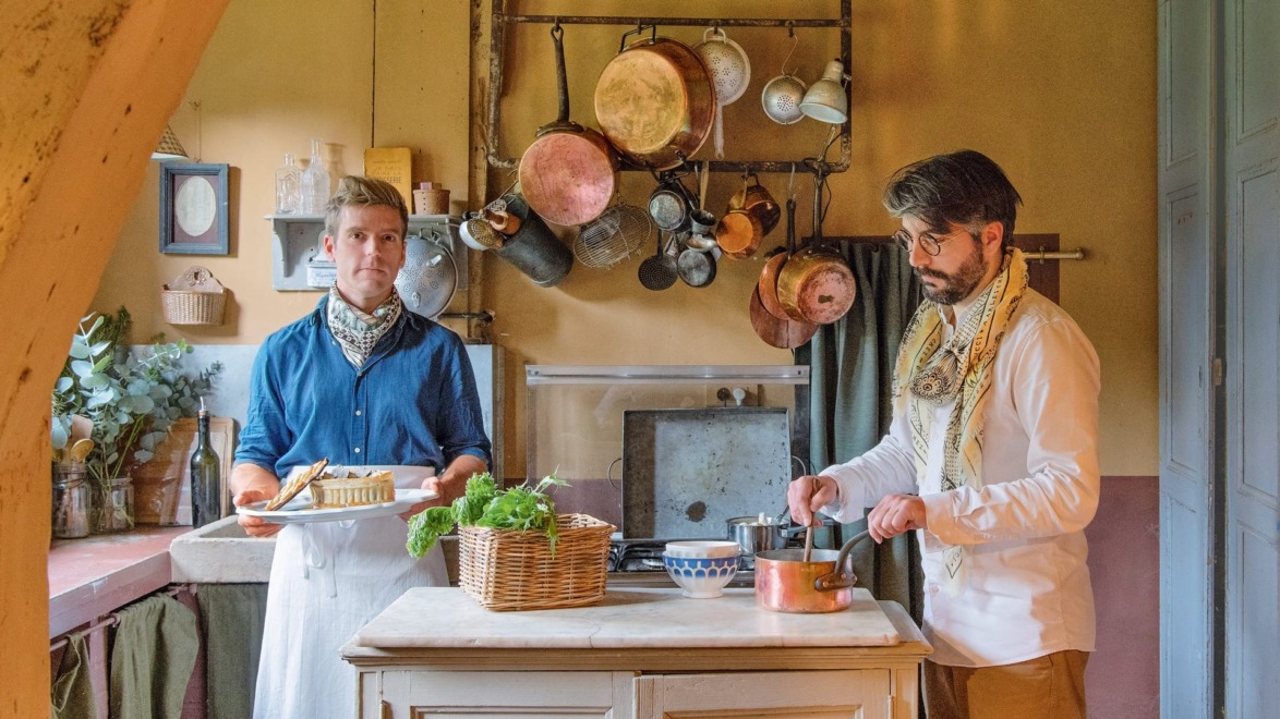 Two men cooking in a rustic kitchen, one holding a pie, the other stirring a pot, with hanging copper cookware.